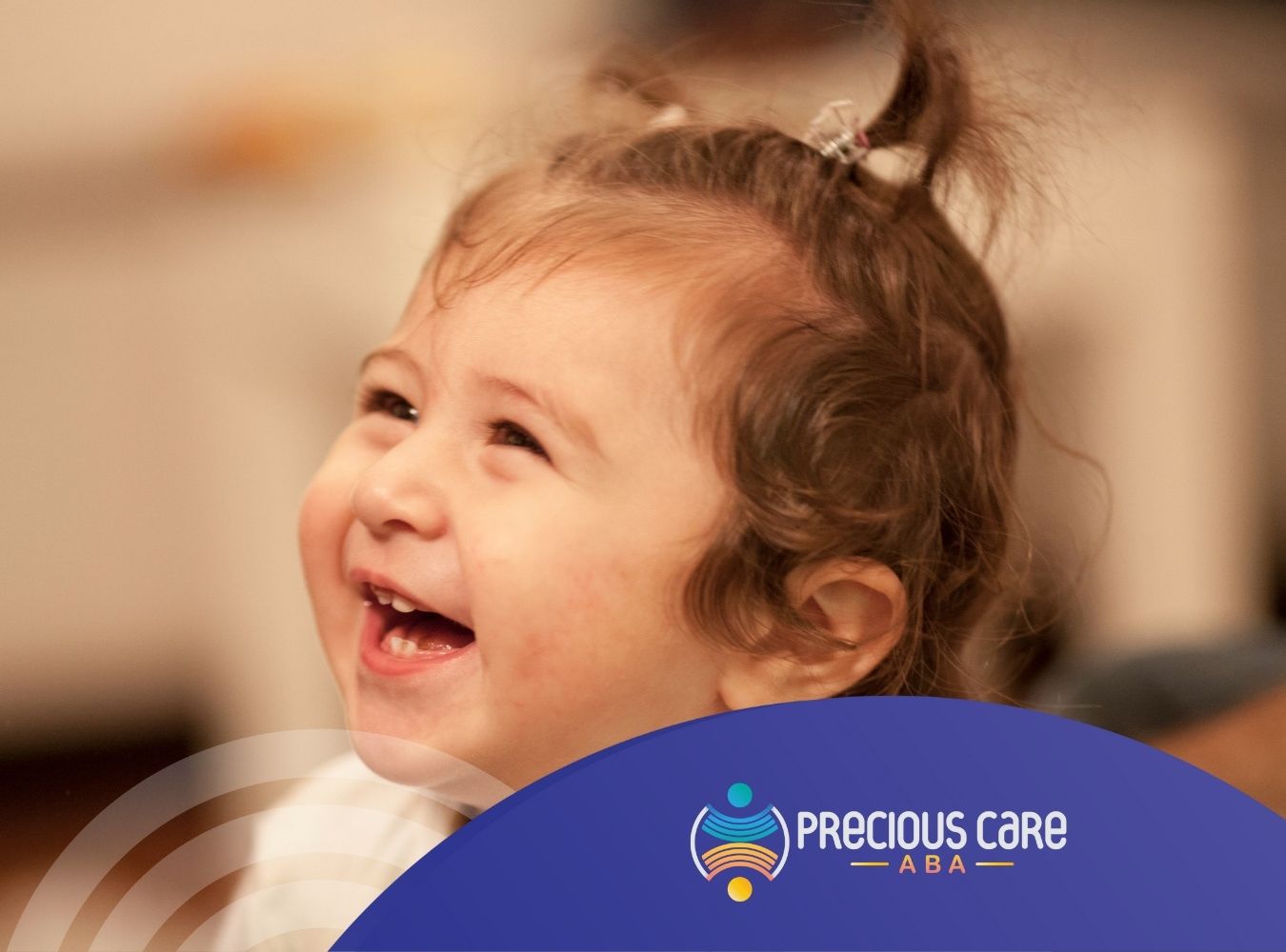 A toddler with curly hair smiles joyfully in a warm indoor setting.