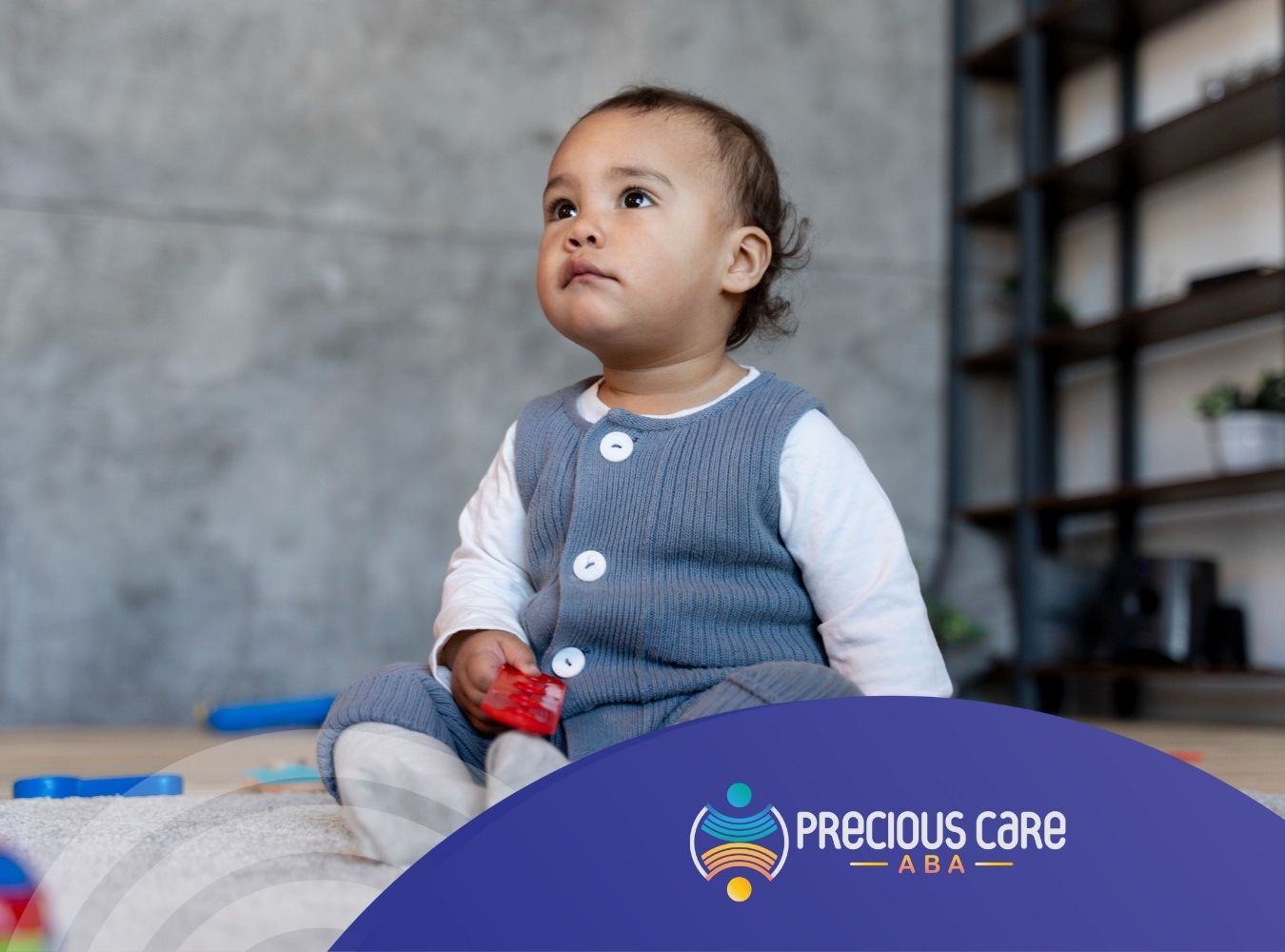 Toddler sitting on a carpet holding a red toy, looking up attentively.
