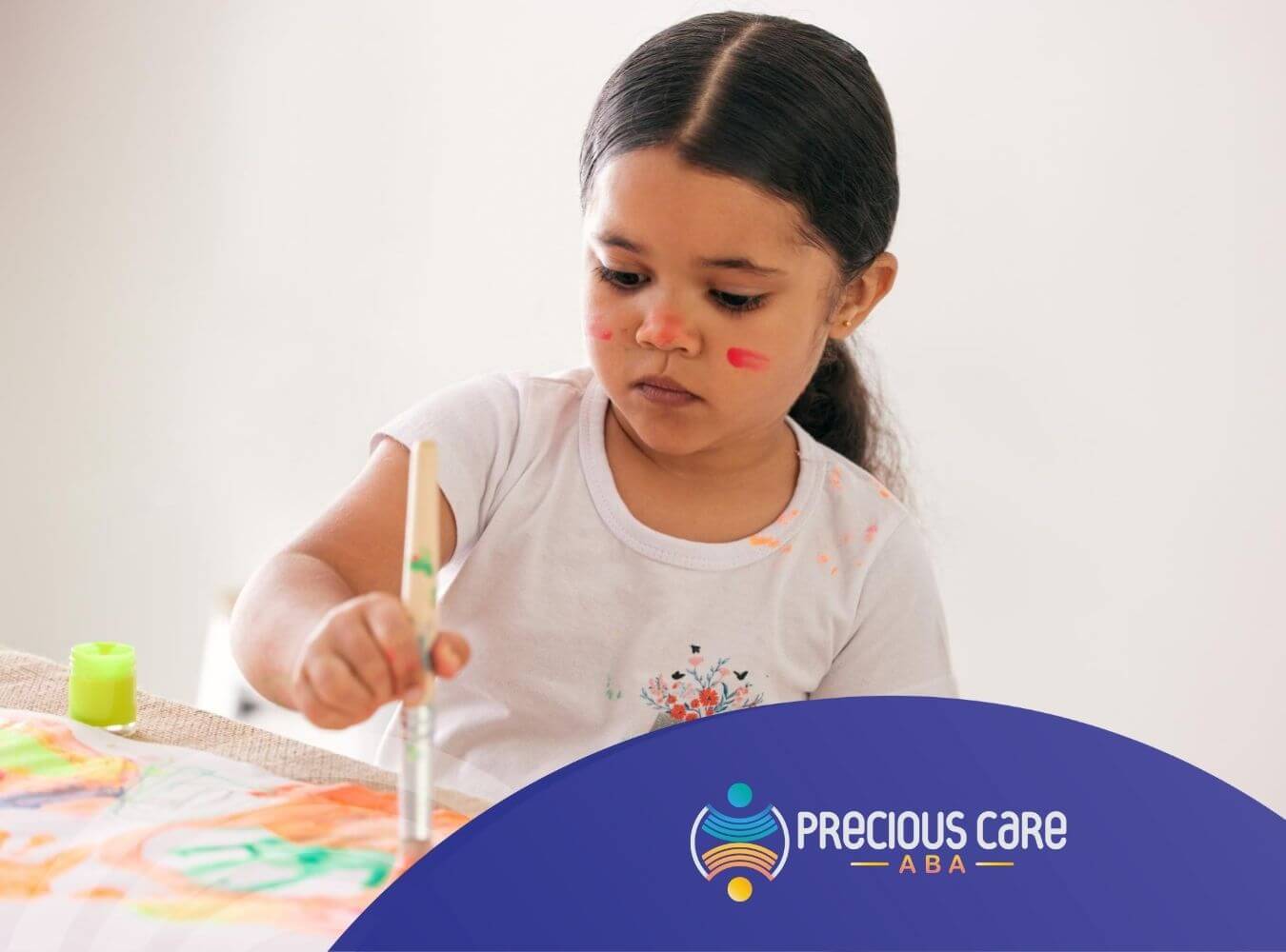 Autistic girl painting with brush and face paint on cheeks during ABA therapy.