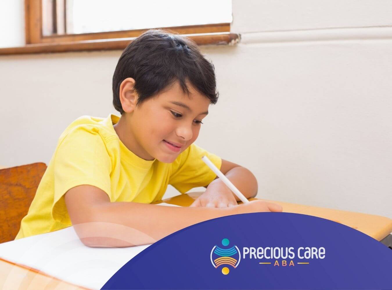 Smiling boy in a yellow shirt writing in a notebook at a school desk.