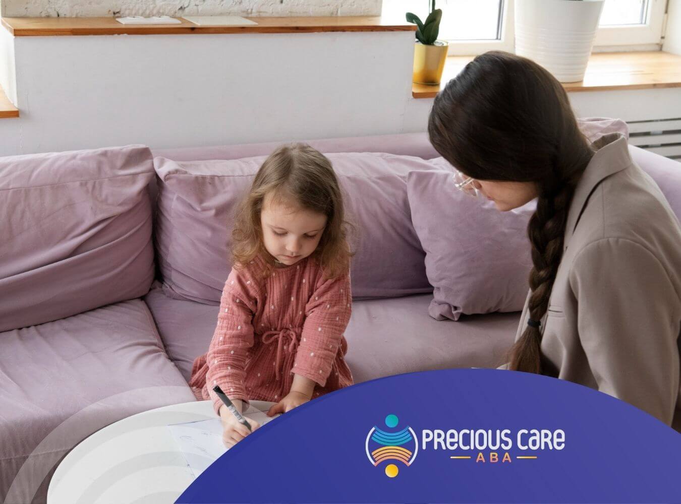Little girl drawing on a clipboard while ABA therapist watches attentively on a couch.