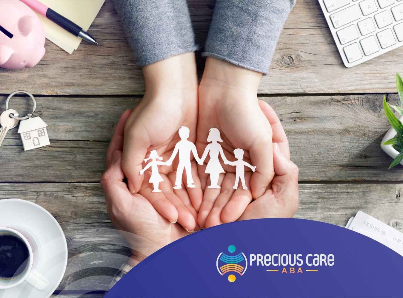 Hands holding paper cutouts of a family on a wooden table with a coffee cup, house key, plant, and keyboard.